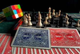 A colorful Rubik's cube sits next to a chessboard with pieces arranged for a game. In the foreground, there are several playing cards with intricate red and blue designs.