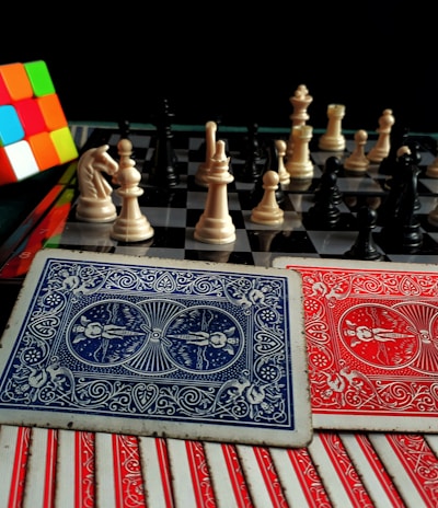 A colorful Rubik's cube sits next to a chessboard with pieces arranged for a game. In the foreground, there are several playing cards with intricate red and blue designs.