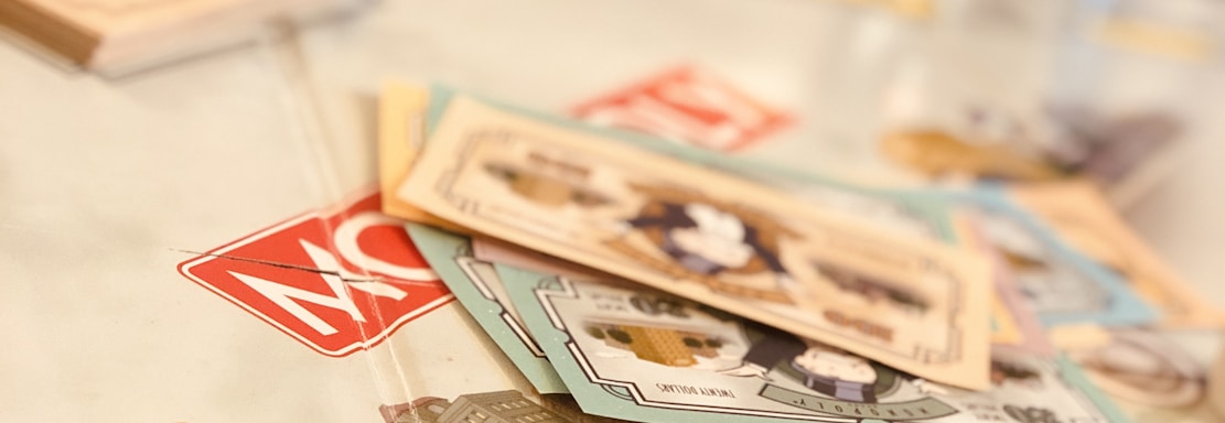 A tabletop setup featuring Monopoly-themed cards and a single die placed on a game board. The cards display various illustrations, while the die shows the number five. The scene is softly lit, creating a warm and inviting atmosphere.