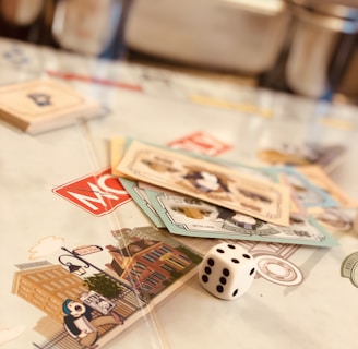 A tabletop setup featuring Monopoly-themed cards and a single die placed on a game board. The cards display various illustrations, while the die shows the number five. The scene is softly lit, creating a warm and inviting atmosphere.