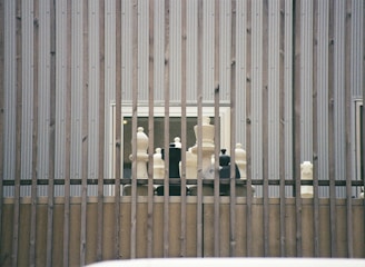 Large chess pieces are arranged on a wooden platform behind vertical metal bars. The setting appears to be outdoors against a corrugated metal wall with two small windows visible.