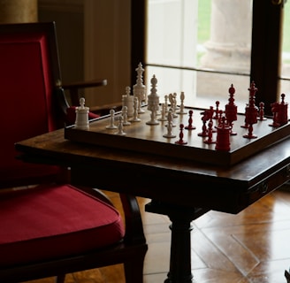 A game of chess with white and red pieces is set up on a wooden board placed on a table. The setup is in a cozy room featuring red cushioned chairs and a large window allowing natural light to enter. The view outside shows a grassy area.