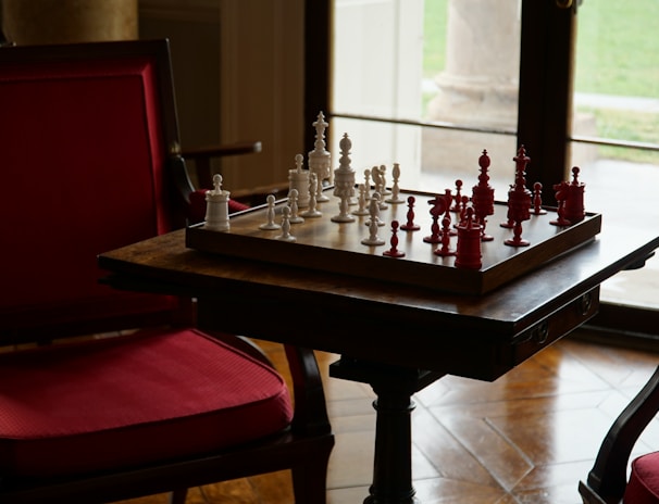 A game of chess with white and red pieces is set up on a wooden board placed on a table. The setup is in a cozy room featuring red cushioned chairs and a large window allowing natural light to enter. The view outside shows a grassy area.