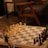 A chessboard set up for a game with white and dark chess pieces positioned for play. The setting includes soft lighting, a wooden table, and comfortable chairs in the background, creating a cozy atmosphere. A small vase with dried flowers is seen to the side of the chessboard.
