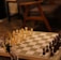 A chessboard set up for a game with white and dark chess pieces positioned for play. The setting includes soft lighting, a wooden table, and comfortable chairs in the background, creating a cozy atmosphere. A small vase with dried flowers is seen to the side of the chessboard.