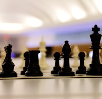 Chess pieces are arranged on a board in a strategic setup, viewed from a low angle. The focus is on the black pieces in the foreground with blurred white pieces in the background. The setting is softly lit with a modern and slightly futuristic atmosphere.