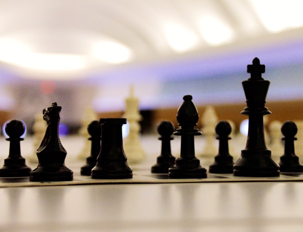 Chess pieces are arranged on a board in a strategic setup, viewed from a low angle. The focus is on the black pieces in the foreground with blurred white pieces in the background. The setting is softly lit with a modern and slightly futuristic atmosphere.