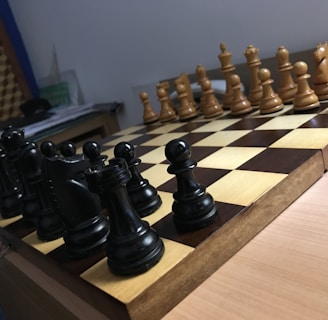 A wooden chessboard is set up for a game with black and white pieces positioned in their starting places. The board is placed on a wooden table, and there is soft lighting in the room. In the background, papers are loosely stacked on a table.