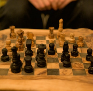 A wooden chessboard with intricately carved chess pieces arranged for a game. The pieces are made of dark and light wood, positioned in a setup suggestive of an ongoing match. A pair of hands is visible in the background, possibly belonging to someone playing the game.