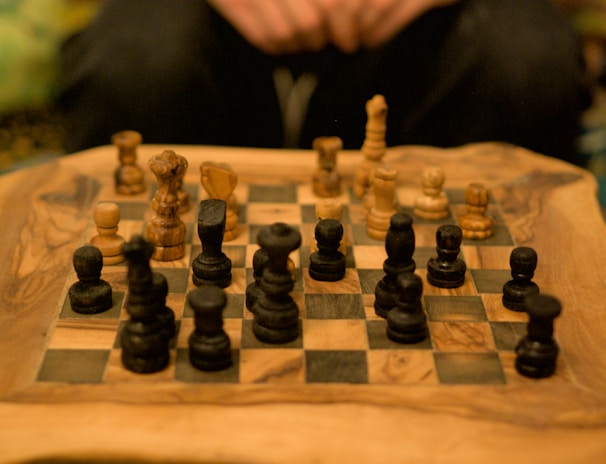 A wooden chessboard with intricately carved chess pieces arranged for a game. The pieces are made of dark and light wood, positioned in a setup suggestive of an ongoing match. A pair of hands is visible in the background, possibly belonging to someone playing the game.
