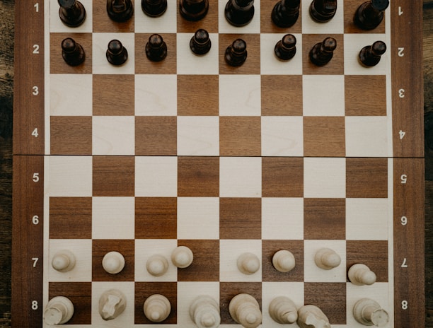 A wooden chessboard is set up with chess pieces in their starting positions. The board is placed on a rustic wooden table, and the chess pieces are made of wood, with darker pieces at the top and lighter pieces at the bottom.