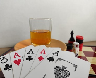 A hand of playing cards featuring four aces and a three of clubs is displayed over a checkered board. A glass of amber liquid rests on an orange saucer in the background. There are stacked poker chips, dice, and a black chess piece nearby.