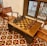 A wooden chessboard set is positioned on a matching wooden table, surrounded by wooden chairs with a classic design. The floor features a decorative, geometric tile pattern with red, white, and black colors. Nearby, a window with vertical bars allows a view of the outside.