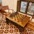 A wooden chessboard set is positioned on a matching wooden table, surrounded by wooden chairs with a classic design. The floor features a decorative, geometric tile pattern with red, white, and black colors. Nearby, a window with vertical bars allows a view of the outside.