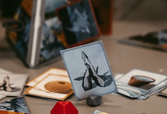 A tabletop game setup with various illustrated cards displayed on a surface. A red, geometric game piece is positioned next to the cards, adding a pop of color. The cards have images and symbols, suggesting a fantasy or strategy theme. The background shows a partially obscured game box with text.
