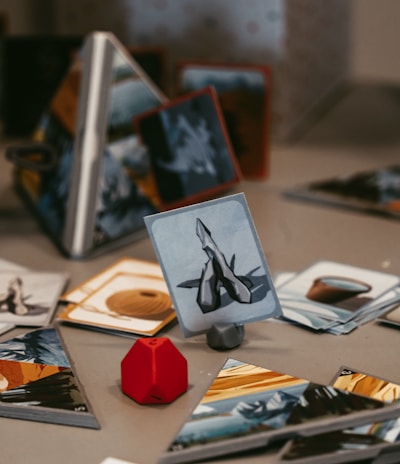 A tabletop game setup with various illustrated cards displayed on a surface. A red, geometric game piece is positioned next to the cards, adding a pop of color. The cards have images and symbols, suggesting a fantasy or strategy theme. The background shows a partially obscured game box with text.