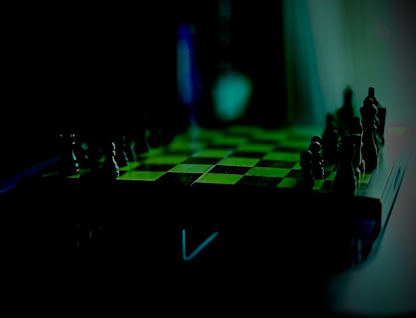 A dimly lit chess board with pieces set up for a game. The focus is on the board, which features a checkerboard pattern. The lighting casts shadows and gives the setting a mysterious effect, highlighting the pieces' silhouettes.
