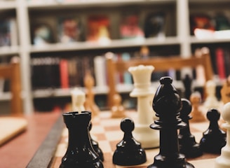 A close-up of a chessboard with black and white chess pieces arranged for play. The background consists of bookshelves filled with various books, creating a cozy, intellectual atmosphere.